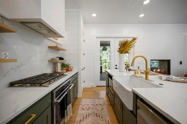 kitchen with sink, stainless steel appliances, light stone counters, exhaust hood, and light wood-type flooring