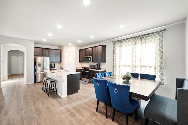 kitchen featuring arched walkways, dark brown cabinetry, a sink, appliances with stainless steel finishes, and an island with sink