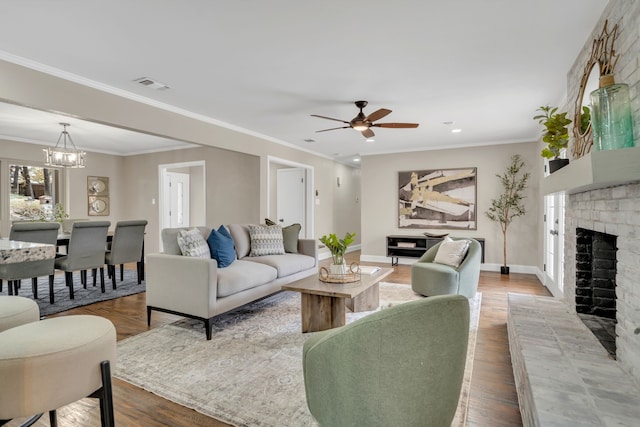 living room with ceiling fan with notable chandelier, wood-type flooring, ornamental molding, and a brick fireplace