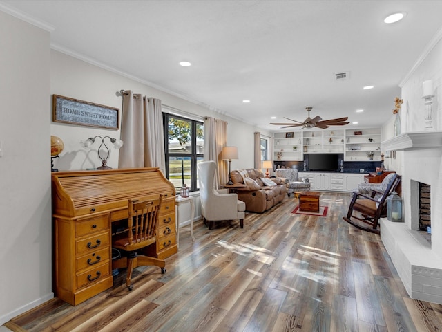 living room with built in features, wood-type flooring, ornamental molding, ceiling fan, and a brick fireplace