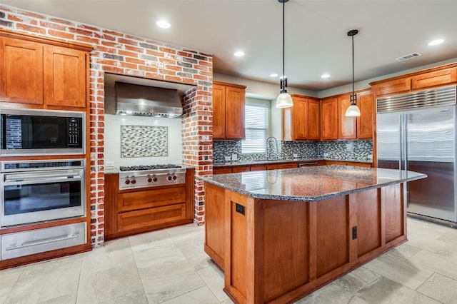 kitchen with decorative light fixtures, dark stone countertops, wall chimney range hood, a kitchen island, and built in appliances