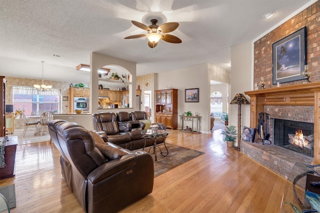 living room with ceiling fan with notable chandelier, a textured ceiling, light hardwood / wood-style floors, and a brick fireplace