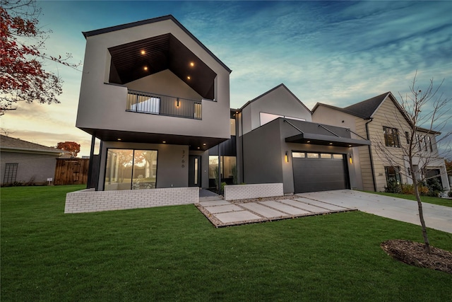 back of property at dusk featuring concrete driveway, fence, a lawn, a balcony, and stucco siding