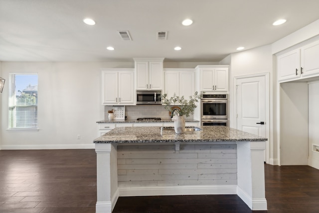 kitchen with dark wood-type flooring, a kitchen island with sink, dark stone counters, appliances with stainless steel finishes, and white cabinetry