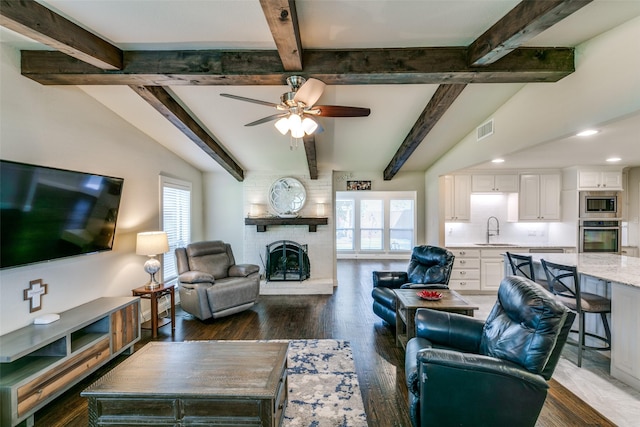 living room featuring ceiling fan, sink, dark wood-type flooring, a brick fireplace, and vaulted ceiling with beams