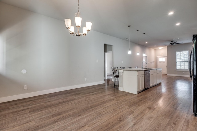 kitchen with pendant lighting, hardwood / wood-style floors, ceiling fan with notable chandelier, stainless steel dishwasher, and an island with sink