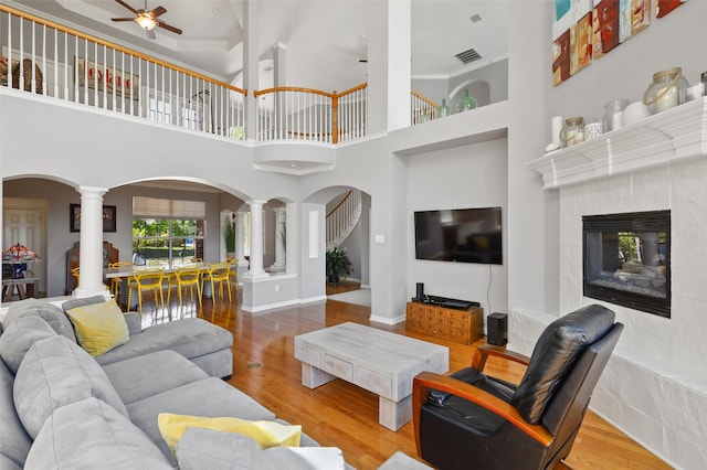 living room with ceiling fan, light hardwood / wood-style flooring, a high end fireplace, decorative columns, and a towering ceiling