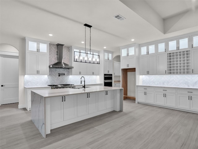 kitchen with white cabinets, decorative light fixtures, wall chimney range hood, and an island with sink