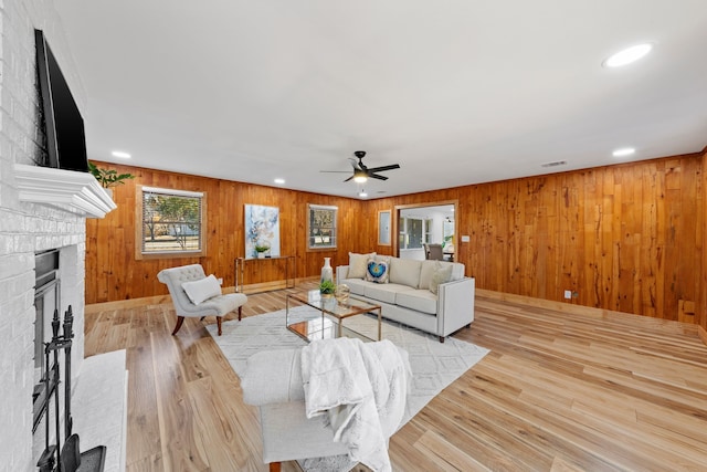 living room featuring wood walls, ceiling fan, light hardwood / wood-style floors, and a brick fireplace