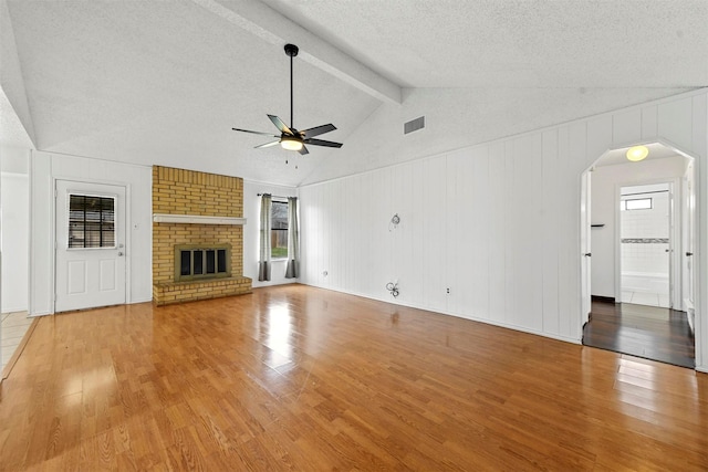 unfurnished living room with ceiling fan, a brick fireplace, lofted ceiling with beams, wood-type flooring, and a textured ceiling