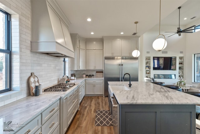 kitchen with light stone countertops, sink, a center island with sink, custom range hood, and appliances with stainless steel finishes