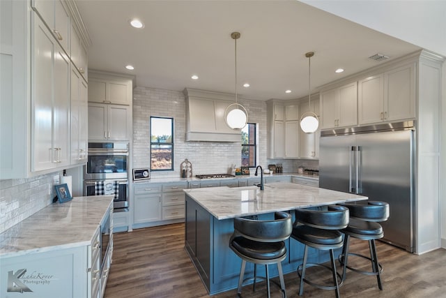 kitchen featuring premium range hood, a kitchen island with sink, dark wood-type flooring, appliances with stainless steel finishes, and decorative light fixtures