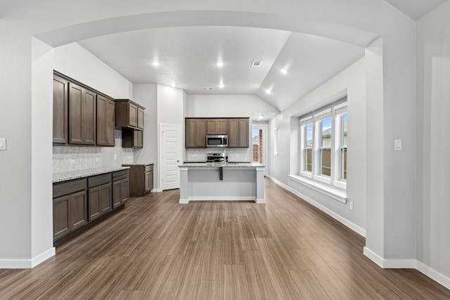 kitchen featuring appliances with stainless steel finishes, backsplash, light stone counters, an island with sink, and dark hardwood / wood-style flooring