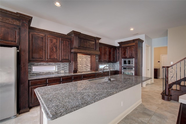 kitchen with dark brown cabinetry, appliances with stainless steel finishes, a sink, dark stone countertops, and backsplash