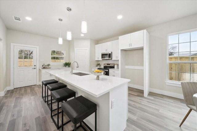 kitchen featuring a kitchen island with sink, sink, appliances with stainless steel finishes, decorative light fixtures, and white cabinetry