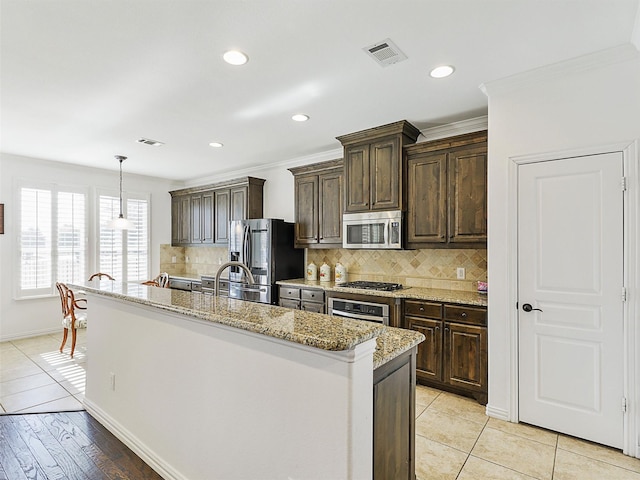 kitchen with appliances with stainless steel finishes, light wood-type flooring, ornamental molding, decorative light fixtures, and an island with sink