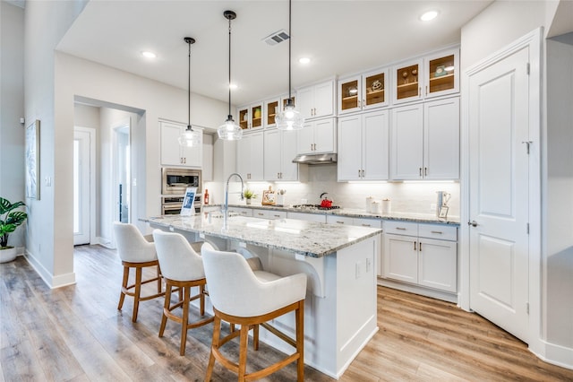 kitchen with appliances with stainless steel finishes, white cabinetry, hanging light fixtures, light stone countertops, and an island with sink