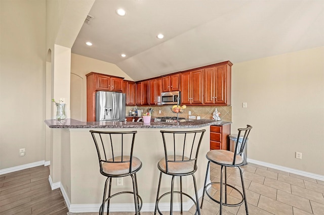 kitchen featuring decorative backsplash, appliances with stainless steel finishes, dark stone countertops, lofted ceiling, and a breakfast bar area