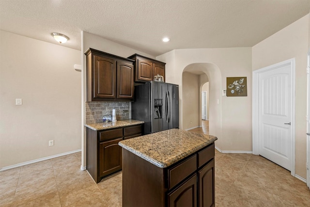 kitchen with dark brown cabinetry, light stone countertops, stainless steel fridge with ice dispenser, backsplash, and a kitchen island