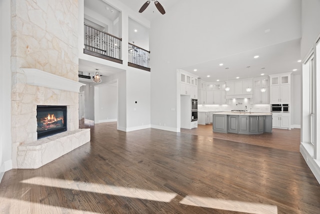 unfurnished living room featuring a high ceiling, a stone fireplace, ceiling fan with notable chandelier, and dark hardwood / wood-style flooring