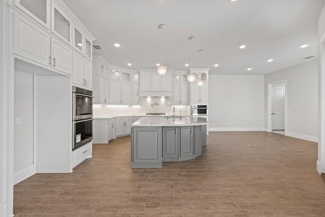 kitchen featuring white cabinetry, tasteful backsplash, hanging light fixtures, double oven, and a kitchen island with sink