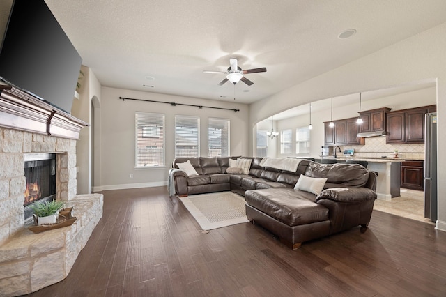 living room with arched walkways, baseboards, dark wood-style floors, ceiling fan, and a stone fireplace