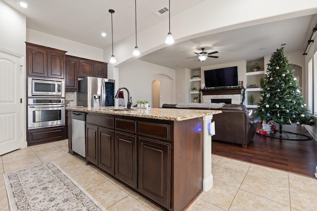 kitchen with appliances with stainless steel finishes, decorative light fixtures, an island with sink, light tile patterned floors, and dark brown cabinets