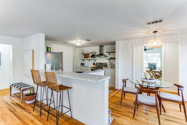 kitchen featuring hanging light fixtures, wall chimney exhaust hood, light hardwood / wood-style floors, kitchen peninsula, and stainless steel appliances