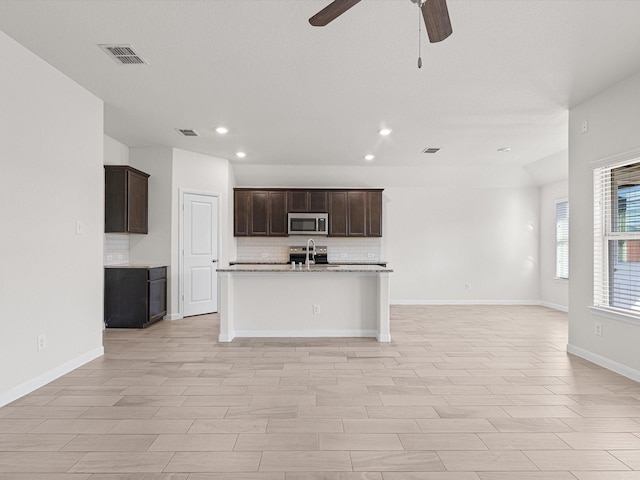 kitchen featuring sink, ceiling fan, decorative backsplash, light stone countertops, and an island with sink