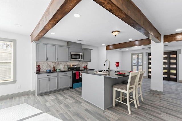 kitchen featuring beamed ceiling, gray cabinets, a center island with sink, and appliances with stainless steel finishes