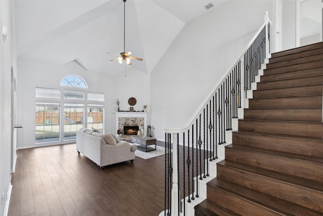 living room with ceiling fan, wood-type flooring, a stone fireplace, and high vaulted ceiling