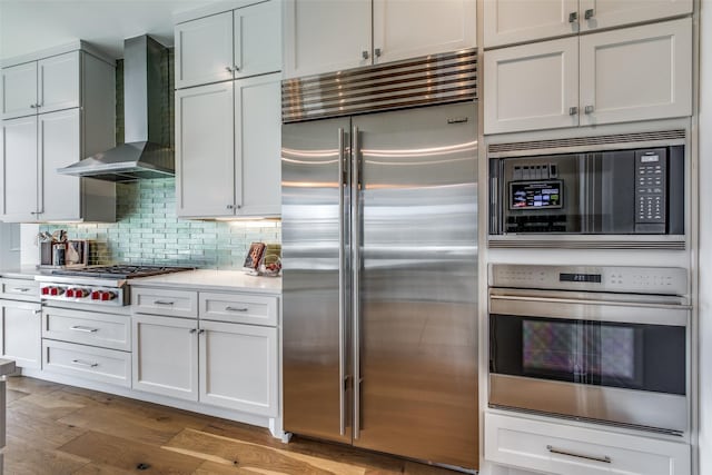 kitchen with built in appliances, light countertops, decorative backsplash, wall chimney exhaust hood, and light wood finished floors