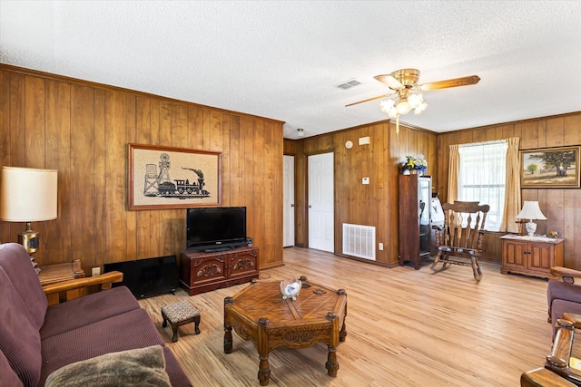 living room with a textured ceiling, light wood-type flooring, ceiling fan, and wood walls