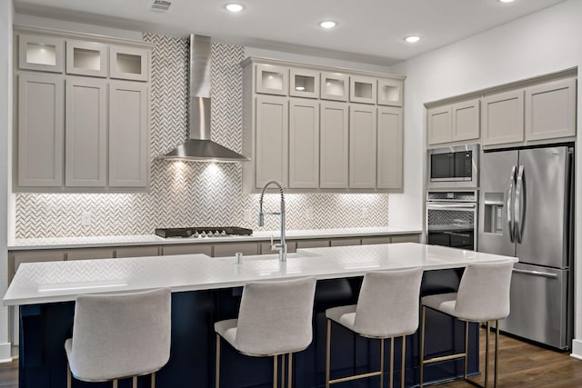 kitchen featuring appliances with stainless steel finishes, dark hardwood / wood-style flooring, tasteful backsplash, wall chimney exhaust hood, and an island with sink