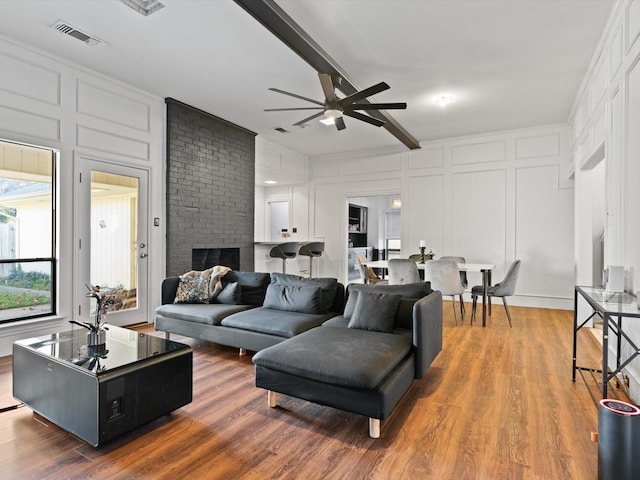 living room with beamed ceiling, wood-type flooring, a brick fireplace, and ceiling fan