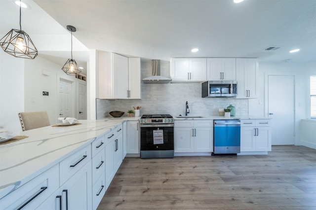 kitchen with white cabinetry, hanging light fixtures, stainless steel appliances, wall chimney range hood, and light hardwood / wood-style flooring