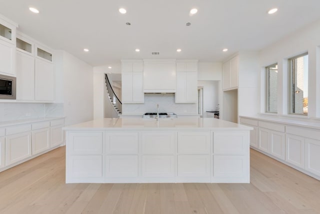 kitchen with white cabinetry, decorative backsplash, light hardwood / wood-style floors, custom range hood, and a center island with sink