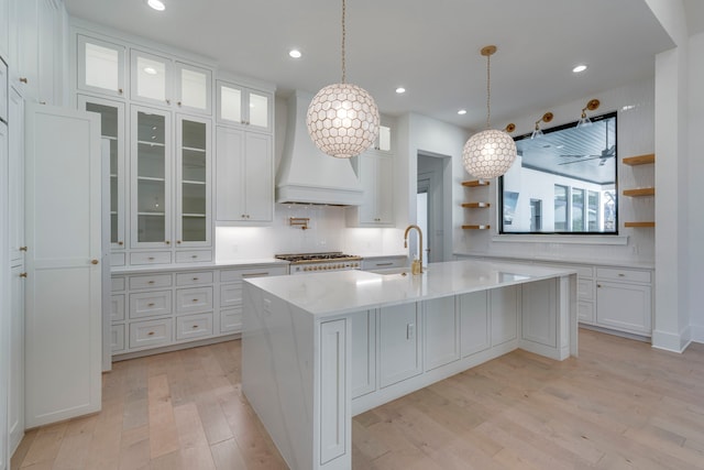 kitchen featuring custom exhaust hood, white cabinetry, decorative light fixtures, light wood-type flooring, and a kitchen island with sink