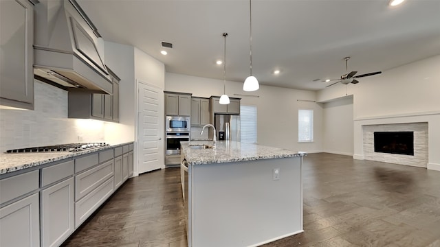 kitchen featuring appliances with stainless steel finishes, gray cabinetry, hanging light fixtures, light stone counters, and a center island with sink