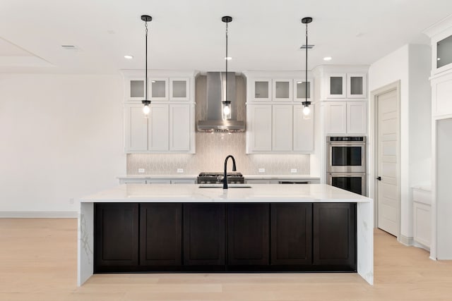 kitchen with double oven, tasteful backsplash, white cabinetry, a kitchen island with sink, and wall chimney exhaust hood