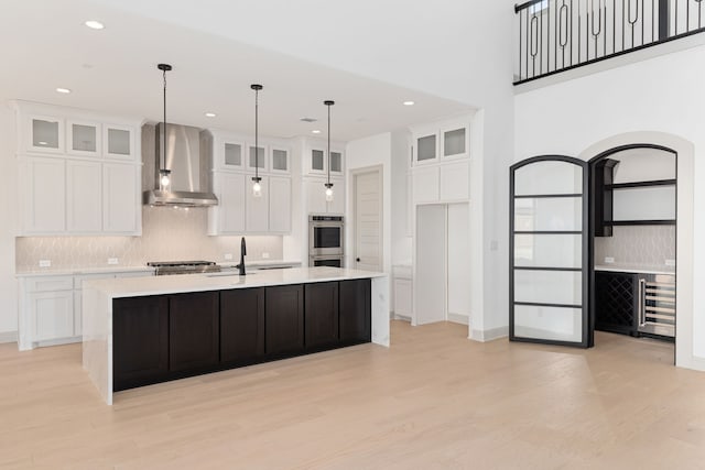 kitchen featuring a center island with sink, wall chimney range hood, white cabinets, and double oven