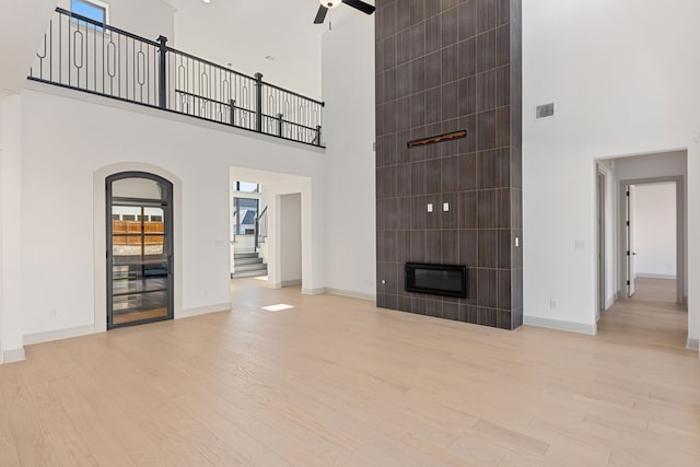 unfurnished living room featuring ceiling fan, a towering ceiling, a tiled fireplace, and light wood-type flooring