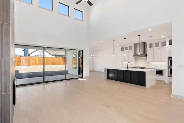 kitchen with decorative light fixtures, white cabinetry, a kitchen island with sink, wall chimney range hood, and light wood-type flooring