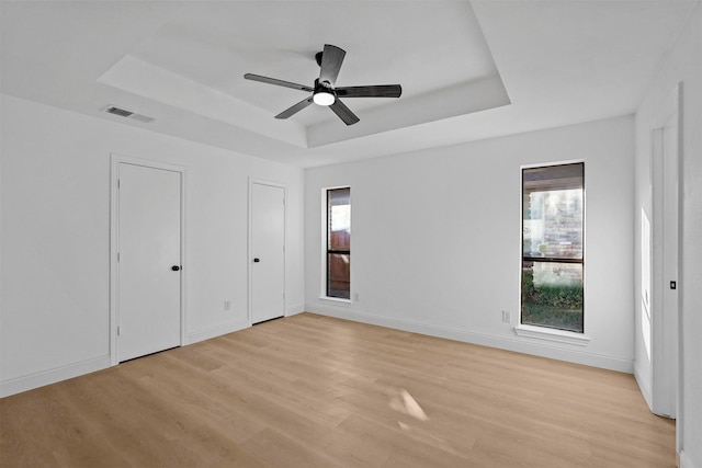 unfurnished bedroom with a tray ceiling, ceiling fan, two closets, and light wood-type flooring