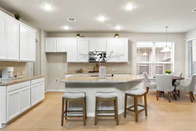 kitchen with pendant lighting, a kitchen island with sink, white cabinets, light wood-type flooring, and light stone counters