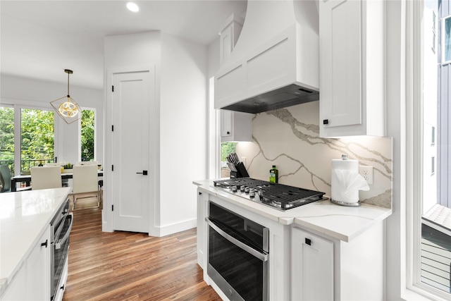 kitchen featuring custom exhaust hood, hanging light fixtures, light wood-type flooring, white cabinetry, and stainless steel appliances