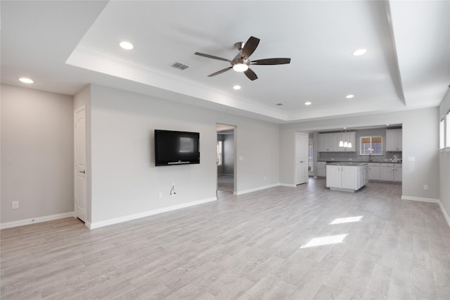 unfurnished living room with a raised ceiling, ceiling fan, sink, and light wood-type flooring