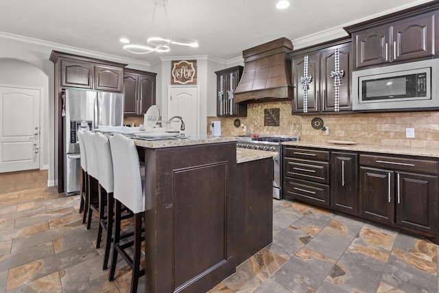 kitchen featuring dark brown cabinetry, an island with sink, stainless steel appliances, and custom range hood