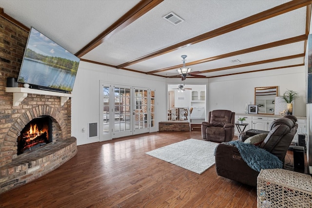 living room with a textured ceiling, wood-type flooring, beamed ceiling, ceiling fan, and a brick fireplace