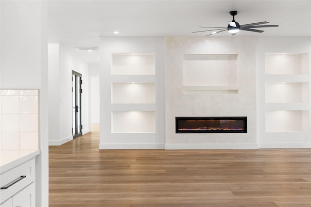 unfurnished living room with ceiling fan, built in shelves, light wood-style flooring, baseboards, and a glass covered fireplace